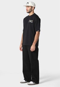 Černé tričko s krátkým rukávem s barevně vyšívanými grafikami, spárováno se volnými černými kalhotami a šedými slip-on botami. Bílé čepice s logem.