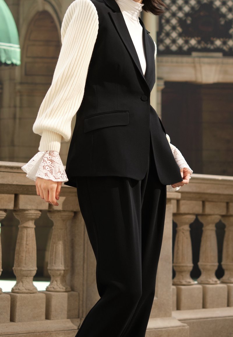 Personne portant un gilet noir sur mesure et un pantalon assorti, par-dessus un col roulé blanc côtelé aux manches bordées de dentelle, se tenant près d'une balustrade en pierre.