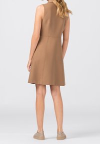 Donna con capelli biondi che indossa un vestito senza maniche color beige fino al ginocchio e sneaker beige, in piedi contro uno sfondo chiaro e uniforme.