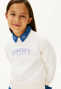 Felpa bianca con logo "TOMMY HILFIGER" viola, abbinata a un colletto in denim blu con dettagli ricamati floreali. Texture liscia.