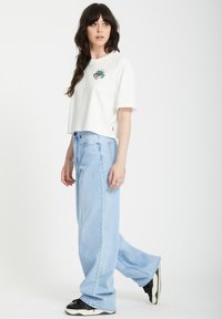 T-shirt blanche coupée avec un design brodé sur la poitrine, associée à un jean large bleu clair et des baskets noires, debout sur un fond uni.