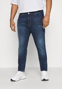 Tmavě modré džíny z denimu s úzkým střihem, pět kapes a žluté švy jako akcenty. Nošeny s bílými sportovními teniskami.