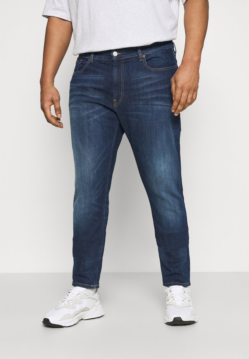 Tmavě modré džíny z denimu s úzkým střihem, pět kapes a žluté švy jako akcenty. Nošeny s bílými sportovními teniskami.