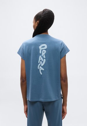 Persona con cabello oscuro trenzado, vistiendo una camiseta y pantalones azules, de espaldas, con texto vertical en azul claro en la parte trasera de la camiseta.