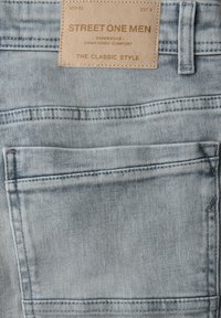 Lichtblauwe spijkerbroek achterzak met een beige leren merkembleem met de tekst "Street One Men The Classic Style."