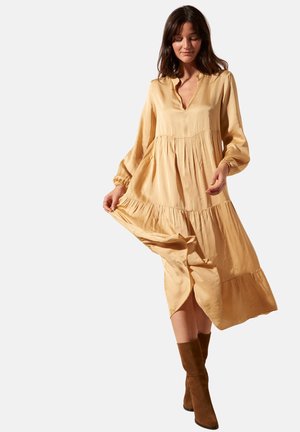 Robe longue beige à volants, en tissu soyeux, avec un décolleté en V, des manches ballon et un ourlet fluide. Portée avec des bottes marron jusqu'aux genoux.