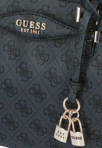 Sac à main en tissu noir avec un motif de logo embossé, accents en matériel doré et un charme à double verrou avec les gravures "EST 1981" et "GUESS".