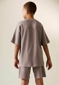 Svetlo šedý, textúrovaný pletený set trička a šortiek, s voľným strihom a krátkymi rukávmi, zobrazený zozadu. Jednoduchý a moderný dizajn.