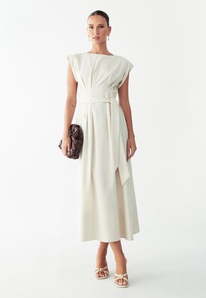 Vrouw draagt een mouwloze beige midijurk met een gestrikte tailleband, houdt een donker gevlochten clutch vast en draagt witte sandalen met hakken en oorbellen.