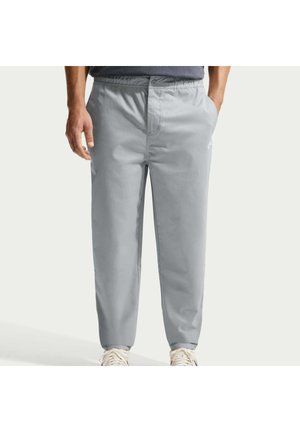 Mand iført lysegrå afslappede bukser med elastisk talje og knap, kombineret med hvide sneakers på en ensfarvet baggrund.