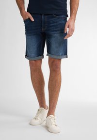 Shorts de denim de color azul oscuro con pu�ños enrollados, que cuentan con bolsillos frontales y un ajuste clásico. Llevados con zapatillas blancas sobre un fondo neutro.