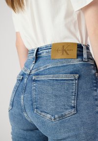 Aukštai pažeminta mėlyna džinsai, pagaminti iš džinsinio audinio su šviesesne skalbine. Yra šviesiai rudos odos etiketė, ant kurios užrašyta "Calvin Klein Jeans", ir įprasti galiniai kišenės.