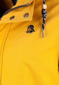 Gul jacka med en slät finish, försedd med en marinblå logotyp av en man i hatt, samt en dragsko med ett träpärla-detalj.