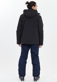 Svart isolerad jacka med huva, ihopparad med marinblå vattentäta byxor. Funktioner inkluderar vida ärmar och dragkedjedetaljer nära manschetterna.