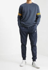 Completo sportivo da uomo con felpa blu marino con strisce gialle e pantaloni da jogging blu marino. Sneakers bianche completano il look.