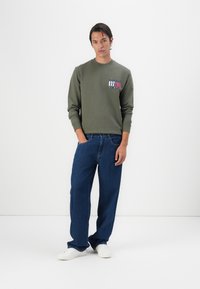 Oliven grøn sweatshirt med et logo-patch på brystet, parret med løstsiddende blå jeans og hvide sneakers. Simpelt design og komfortabel pasform.