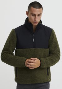 Green fleece pullover with a black nylon upper section and high collar; features a half-zip closure and side pockets. Textured fabric.