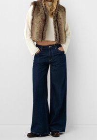 Faux fur vest in brown over a white ribbed crop top, paired with wide-leg dark blue jeans featuring front pockets and a classic button closure.