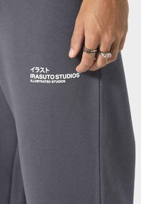 Grå bomulds sweatpants med en glat tekstur. Har hvide trykte logoer: "IRASUTO STUDIOS" og japanske tegn. Hånd med ringe synlig.