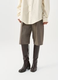 Barna bőr térdfeletti csizma négyzet alakú sarokkal, szürke kockás, térd feletti rövidnadrággal és krém színű inggel viselve.