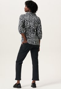 Kvinde med krøllet hår iført en sort- og hvid leopardmønstret bluse, sorte jeans og sorte loafers, stående med den ene hånd i lommen.