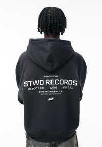 Czarna bluza z kapturem z dużym białym nadrukiem na plecach, zawierającym napis "STWD RECORDS" oraz inne detale. Materiał wydaje się miękki; wyposażona w kaptur.