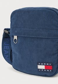 Ναυτικό καπιτονέ τσάντα ώμου με φερμουάρ και τσέπη, με υφασμάτινο λουρί που έχει την ένδειξη "JEANS" και λογότυπο σε κόκκινο, άσπρο και μπλε.