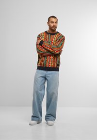 Kleurrijke gebreide sweater met rode, groene en gele geometrische patronen; ribgebreide mouwen en zoom; gecombineerd met lichtblauwe wide-leg jeans en witte schoenen.