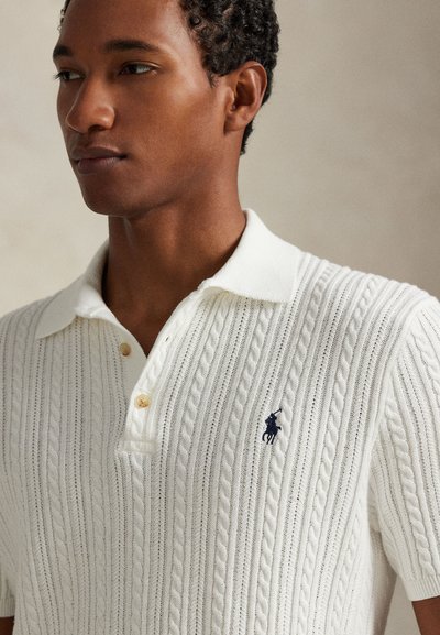 Gros plan sur une personne portant un polo blanc à manches courtes en maille torsadée avec un col et un petit logo brodé foncé sur la poitrine.