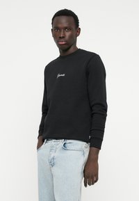 Felpa nera con scollo rotondo e maniche lunghe, caratterizzata da testo ricamato bianco sul petto. Abbinata a jeans di jeans azzurri chiari.