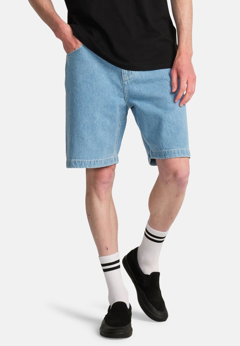 Τζιν σορτς σε ανοιχτό μπλε χρώμα με ίσια γραμμή, πλαϊνές τσέπες και γυρισμένο τελείωμα, συνδυασμένα με μαύρα slip-on παπούτσια και κάλτσες με λευκές ρίγες.