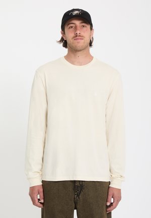 Homme portant une chemise crème à manches longues, un pantalon marron olive et une casquette noire avec un motif de scorpion, debout devant un fond blanc.