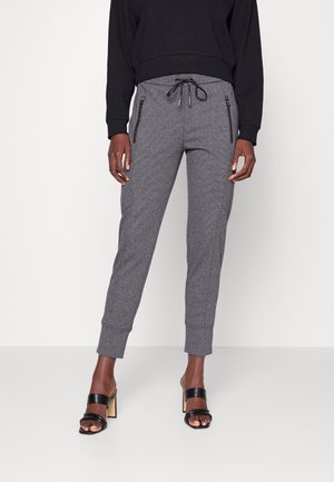 Person wearing gray jogger pants with black zipper pockets, a black sweatshirt, and black high-heeled sandals standing against a white background.