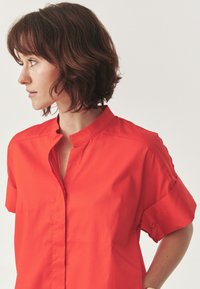 Red short-sleeve blouse with a stand collar, featuring a front placket, smooth texture, and a relaxed fit. No visible patterns or accents.