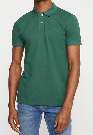 Homme portant un polo vert foncé moulant à manches courtes avec deux boutons blancs et un jean bleu sur fond uni.