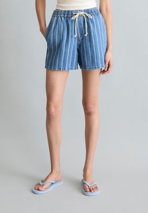 Person trägt blau-weiß gestreifte Jeansshorts mit Kordelzug an der Taille, weißes Tanktop und hellblaue Flip-Flops vor grauem Hintergrund.