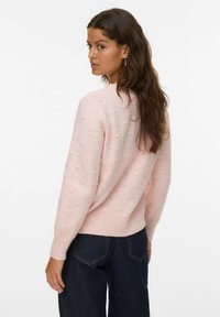 Ženska z dolgimi rjavimi lasmi, oblečena v roza teksturirano pulover in temno modre kavbojke, se obrača čez ramo pred enostavno ozadje.