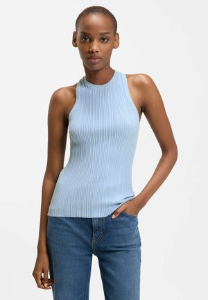 Femme portant un haut sans manches côtelé bleu clair et un jean bleu, debout avec une main dans la poche devant un fond blanc.