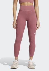 Leggings in staubigem Rosé, hoch geschnitten, dehnbarem Stoff mit tonalen Nähten. Verfügt über ein kleines weißes Logo. Kombiniert mit hellfarbenen Sportschuhen.