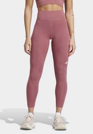 Leggings in staubigem Rosé, hoch geschnitten, dehnbarem Stoff mit tonalen Nähten. Verfügt über ein kleines weißes Logo. Kombiniert mit hellfarbenen Sportschuhen.
