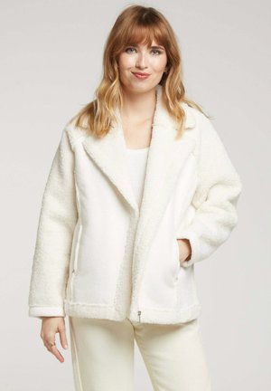 Veste en polaire blanche et duveteuse avec un design à texture double, grands revers et deux poches latérales. Elle possède une coupe droite et une fermeture éclair.