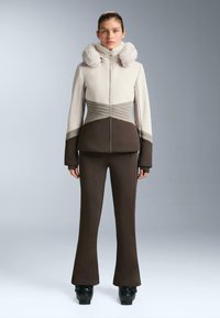 Zweiteiliges Outfit mit einer beigefarbenen Jacke mit Reißverschluss und pelzgefütterter Kapuze sowie braunen ausgestellten Hosen. Die Jacke hat strukturierte Muster und gerippte Details.