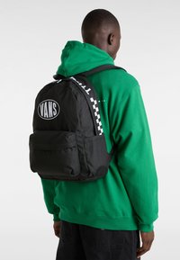 Musta kangaskassi, jossa on ruudullinen hihna, suurella valkoisella "VANS"-logolla ja etutaskulla. Päällä vihreä huppari.