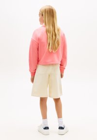 Ροζ φούτερ με χαλαρούς ώμους, μπεζ σορτς μήκους μέχρι το γόνατο, λευκές κάλτσες αστραγάλου και λευκά αθλητικά παπούτσια με ναυτικό μπλε λεπτομέρειες. Απλό φόντο.