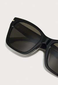 Gafas de sol negras de gran tamaño con lentes oscuros, forma rectangular elegante, acabado brillante, acento metálico en la montura y construcción robusta.