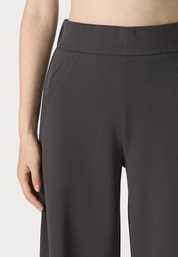 Un pantalon large gris en tissu lisse présente une taille haute et des poches latérales, mettant en valeur un design épuré et sur mesure.