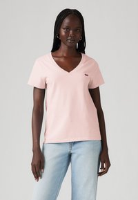 T-shirt rose clair en V en coton, avec des manches courtes et un petit logo de marque sur la poitrine gauche. Porté avec un jean bleu clair.