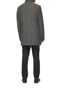 Ein Mann steht mit dem Rücken zur Kamera, trägt einen grauen strukturierten Übermantel, schwarze Hose und schwarze Anzugschuhe vor einem weißen Hintergrund.