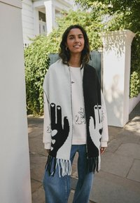 Châle en mélange de laine avec des motifs à main noir et blanc, doté de bords frangés. Le modèle le porte par-dessus un pull léger et un jean bleu.