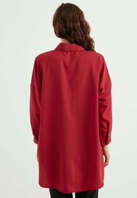 Rotes, übergroßes Hemd mit Kragen, langen Ärmeln, geknöpften Manschetten und einer glatten Textur. Rückansicht zeigt einen leicht geschwungenen Saum.